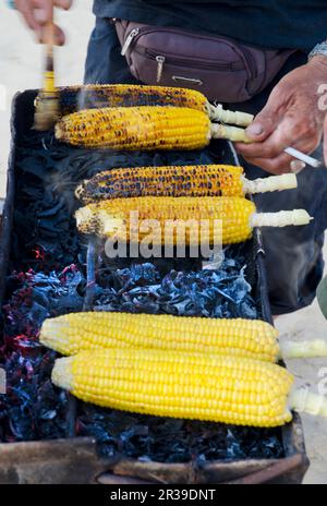 Un vendeur de rue balinais vendant des épis de maïs épicés sur un barbecue au charbon de bois avec une cigarette dans la main Banque D'Images