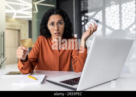 Une femme hispanique choquée et bouleversée travaillant au bureau, une femme d'affaires regardant la caméra tenant une carte de crédit bancaire avec colère, une cliente sur le lieu de travail avec un ordinateur portable a reçu une erreur de paiement en ligne. Banque D'Images