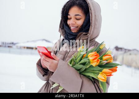 Jeune femme africaine enchanteresse en vêtements chauds et décontractés tient le téléphone et le bouquet de tulipes orange. Joyeuse fille de course mixte avec fleurs de printemps brillant envoyer Banque D'Images