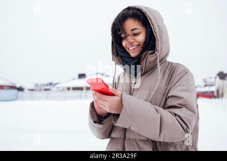 Jeune femme africaine charmante dans des vêtements chauds et décontractés tenant le téléphone rouge dans ses mains. Joyeux sourire fille de course mixte en utilisant le mobile et l'envoi de message. W Banque D'Images