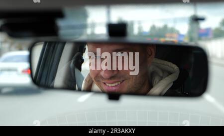 Joyeux homme caucasien adulte conduisant une voiture dans la ville regardant et souriant dans le rétroviseur. Visage d'homme heureux reflété dans le rétroviseur de voiture. Déplacement Banque D'Images