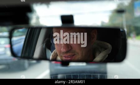 Joyeux jeune homme caucasien conduisant une voiture dans la ville regardant et se clignant dans le rétroviseur. Un homme souriant et charmant se reflète dans le miroir de la voiture. Déplacement Banque D'Images