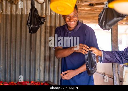 Jeune homme africain excité vendant des tomates dans un marché africain local à un client, recevant de l'argent Banque D'Images