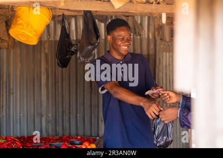 Jeune homme africain excité vendant des tomates dans un marché africain local à un client, recevant de l'argent Banque D'Images