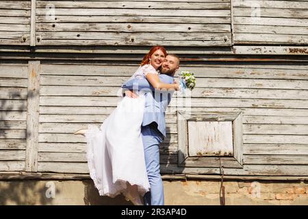 Juste marié aimant hipster couple dans la robe de mariage et le costume posant en face d'une vieille maison en bois. Banque D'Images