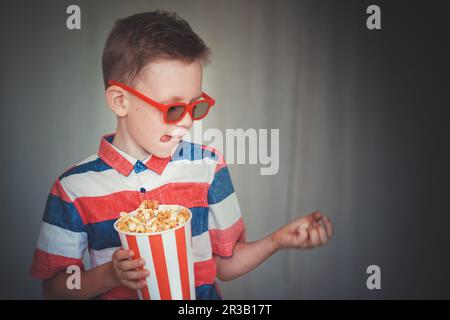 Un jeune garçon regarde un film en 3D verres au cinéma ou à la maison. Un petit enfant mange du pop-corn sur un bac gris Banque D'Images