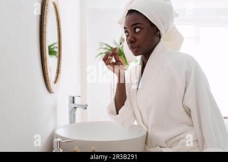 Femme portant un peignoir en utilisant un rouleau de jade sur le visage dans la salle de bains à la maison Banque D'Images