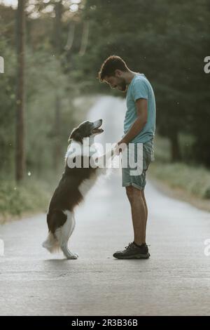 Jeune homme debout avec un chien sur la route au coucher du soleil Banque D'Images