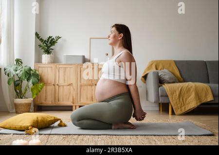 Femme enceinte étirant les mains assis sur le tapis à la maison Banque D'Images