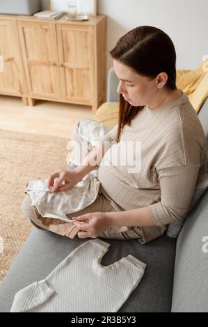 Femme enceinte pliant des vêtements de bébé assis sur un canapé à la maison Banque D'Images