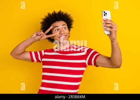 Photo de funky handsome personne tenir le smartphone show v-sign faire selfie isolé sur fond jaune de couleur Banque D'Images