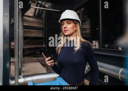 Femme d'affaires réfléchie portant un casque et tenant un smartphone à l'usine Banque D'Images