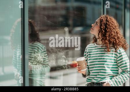 Jeune femme regardant le mur en verre tenant une tasse à café jetable Banque D'Images