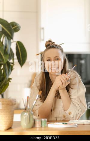 Jeune femme souriante tenant un pinceau sur la table à la maison Banque D'Images