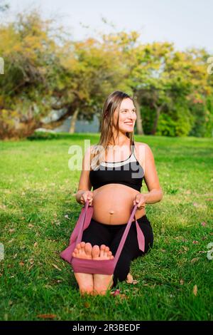 Femme enceinte souriante s'étirant avec un bracelet de résistance sur la pelouse Banque D'Images