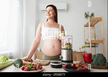 Femme enceinte souriante préparant un smoothie aux fruits dans la cuisine à la maison Banque D'Images