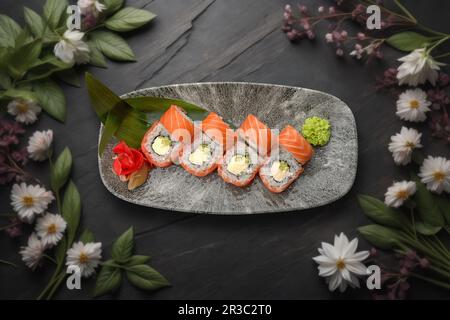 rouleau de sushi au saumon, au fromage crémeux et à l'avocat sur une surface en pierre texturée ornée de décorations florales Banque D'Images