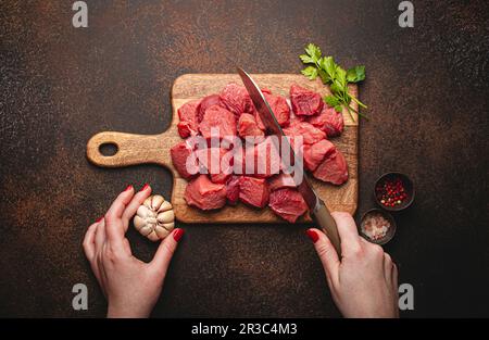 Les mains des femmes hacher de la viande de bœuf crue en cubes sur une planche à découper en bois pour cuire le ragoût Banque D'Images