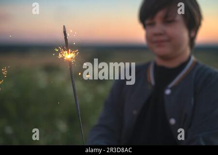 Un adorable garçon tient des spameux dans ses mains, des feux d'artifice dans la soirée en été au coucher du soleil. Ambiance festive. Banque D'Images