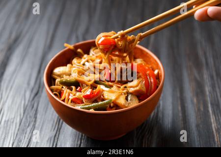 Assiette de nouilles de sarrasin avec légumes, champignons, viande de poulet sur fond sombre. Les bâtonnets chinois prennent le soba japonais d'un bol en argile Banque D'Images