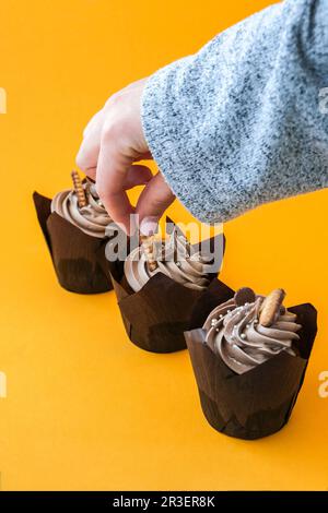 Les mains des femmes font de délicieux petits gâteaux au chocolat avec de la crème sur fond jaune. Trois muffins au chocolat. Fête gâteau d'anniversaire pr Banque D'Images