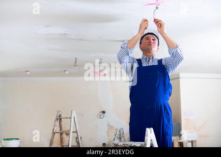 Électricien en combinaison bleue fait le câblage électrique sur le plafond dans la salle en cours de rénovation Banque D'Images