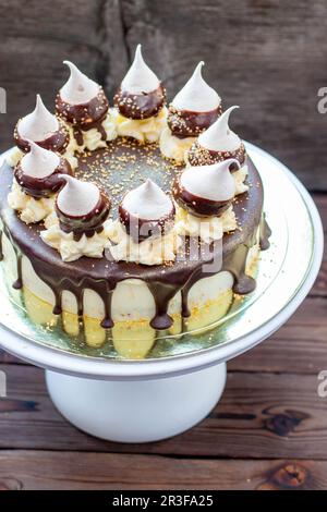 Cheesecake d'anniversaire décoré avec une sauce au chocolat noir fondu, des meringues et des tourbillons de crème au beurre sur le cakestan blanc. Arrière-plan rustique Banque D'Images