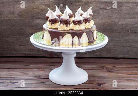 Cheesecake d'anniversaire décoré avec une sauce au chocolat noir fondu, des meringues et des tourbillons de crème au beurre sur le cakestan blanc. Arrière-plan rustique Banque D'Images