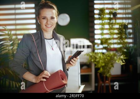 Un lieu de travail durable. femme moderne souriante propriétaire de petite entreprise dans un bureau moderne vert avec casque sans fil, tapis de yoga et smartphone. Banque D'Images