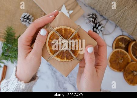 Femme faisant boîte avec les cadeaux du nouvel an, enveloppé dans du papier artisanal et décoré avec des tranches d'orange sèches. Concept de vacances et de cadeaux. Banque D'Images