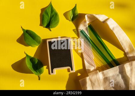 Pailles en verre réutilisables sur fond coloré avec feuilles vertes dans un sac écologique écologique. Modèle de maquette de tableau noir vide pour cop Banque D'Images