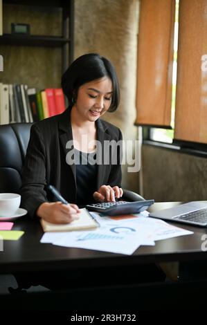 Une femme d'affaires asiatique ou une femme entrepreneure d'affaires, belle et prospère, calcule les ventes à l'aide d'une calculatrice et d'un ordinateur portable à son bureau Banque D'Images