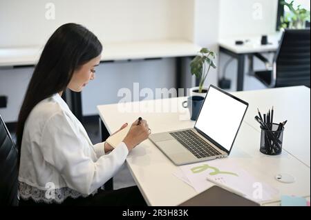 Vue latérale d'une belle femme d'affaires asiatique prenant des notes sur son carnet personnel, travaillant à son bureau dans le bureau. ordinateur portable écran blanc mo Banque D'Images