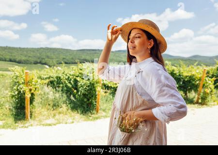 Jeune belle femme en chemise blanche, beige sundress et chapeau de paille regarde le ciel de rêve. Une brune attrayante sourit sur fond de vignobles et Banque D'Images