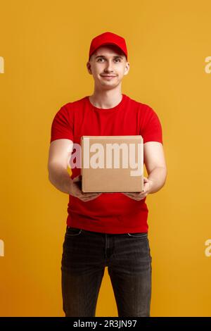 Employé de livreur dans un capuchon rouge, t-shirt rouge tenant une boîte en carton isolée sur fond jaune Banque D'Images