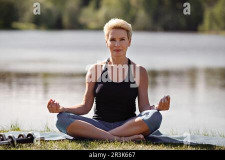 Calme femme mature pratique le yoga à l'extérieur. Athlète féminine assise en position lotus sur fond d'eau de la nature. Mode de vie actif et santé. Banque D'Images