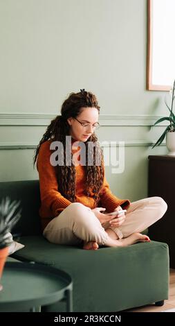 Jeune femme se détendant et magasiner en ligne avec un smartphone à la maison. Bonne dame appréciant les médias sociaux sur téléphone mobile à la maison. Femme attrayante utilisant Banque D'Images