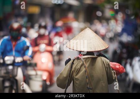 Accent sélectif sur le chapeau conique traditionnel de personne marchant contre les motos de circulation sur la rue animée dans le Vieux quartier à Hanoi, Vietnam. Banque D'Images