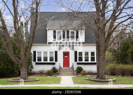 Une belle maison blanche, suburbaine avec une porte d'entrée rouge et de grands arbres dans la cour avant. Banque D'Images
