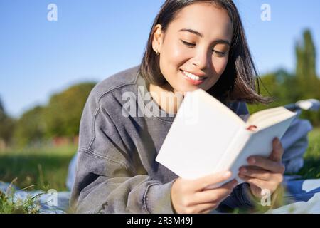 Portrait de belle fille asiatique souriante, lisant dans le parc, allongé sur l'herbe avec le livre préféré. Concept loisirs et personnes Banque D'Images