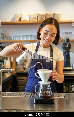 Plan vertical de barman asiatique souriant, barista en tablier bleu, verser de l'eau dans une petite bouilloire, préparer du café derrière le comptoir Banque D'Images