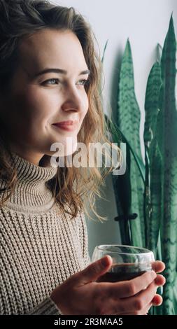 Une jeune femme sereine savoure une tasse de thé chaud tout en admirant le paysage urbain époustouflant d'une fenêtre, en prenant un moment paisible pour se détendre et Banque D'Images