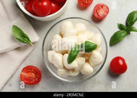 Délicieuses boules de mozzarella dans un bol en verre, tomates et feuilles de basilic sur une table gris clair, à plat Banque D'Images