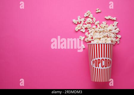 Gobelet en papier renversé avec délicieux pop-corn sur fond rose, plat. Espace pour le texte Banque D'Images