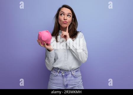 adorable femme brune de 25 ans dans une chemise et un jean décide de dépenser son épargne d'une banque de porc Banque D'Images