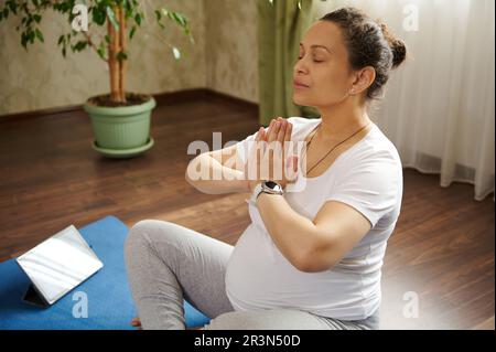 Magnifique femme enceinte garde les mains paumes ensemble, faisant le yoga de grossesse et exercices de souffle à la maison. Cours de yoga en ligne Banque D'Images