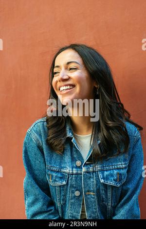 Portrait d'une jeune femme hispanique heureuse aux cheveux foncés et en veste en denim souriant tout en regardant loin et debout sur fond orange Banque D'Images