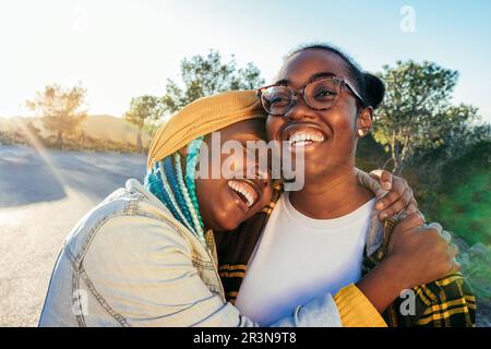 Riant la femme afro-américaine avec les yeux fermés embrassant regardant loin ami ethnique dans des lunettes tout en se tenant ensemble sur la route contre le vert tre Banque D'Images