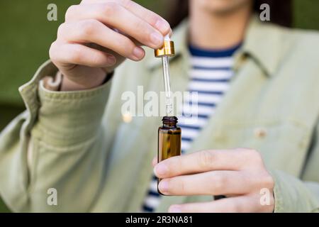 Anonyme femme dans des vêtements décontractés qui goutte l'huile liquide du tube dans une petite bouteille tout en se tenant sur un fond flou Banque D'Images