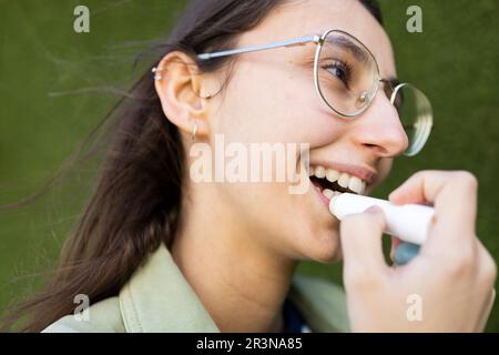 De dessous de jeune femme en lunettes souriant et appliquant le baume à lèvres tout en regardant loin sur fond vert Banque D'Images